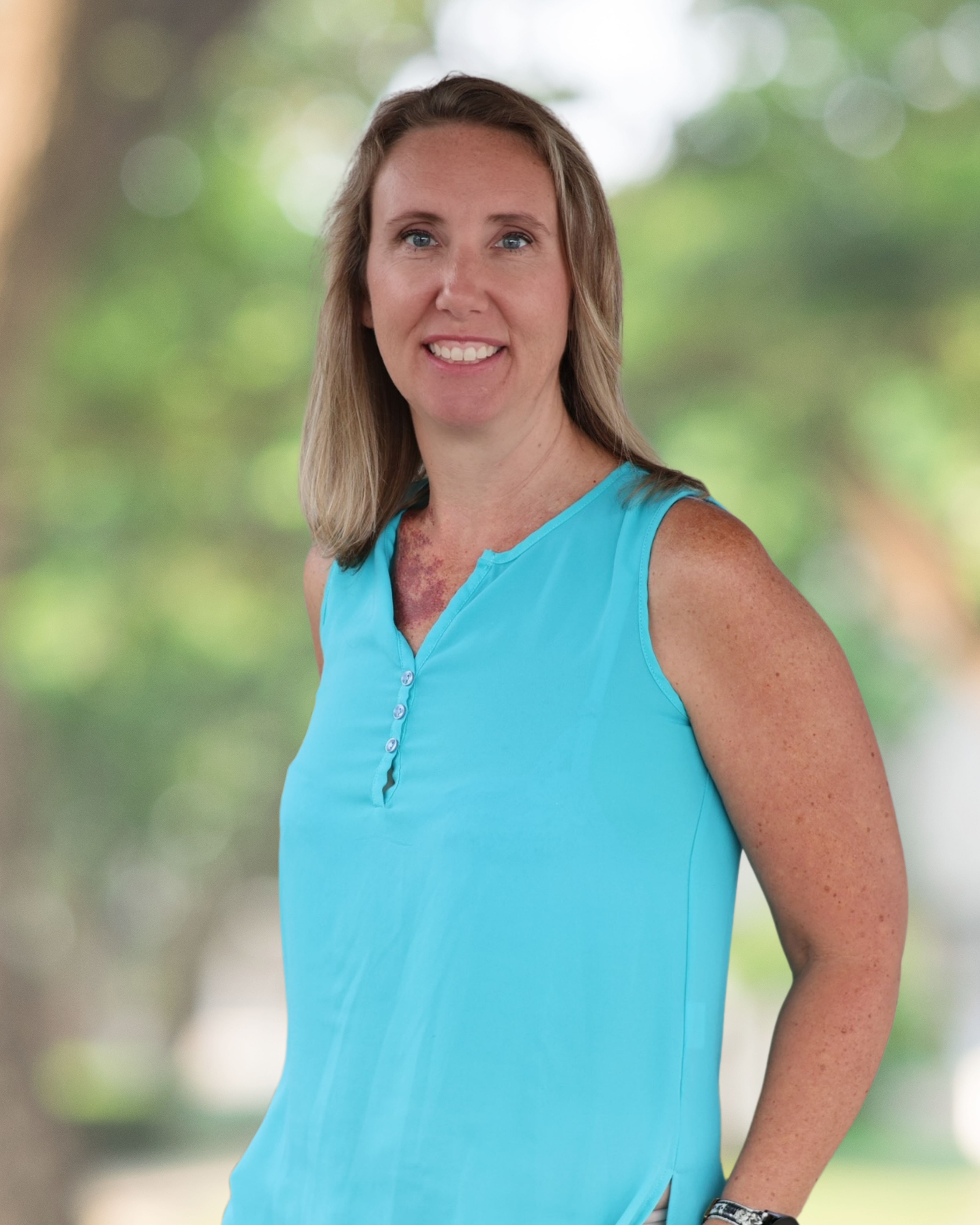 Professional headshot of a smiling woman standing outdoors against a softly blurred background of green trees. She is wearing a sleeveless turquoise blouse and has shoulder-length light brown hair. She faces the camera with a friendly expression.