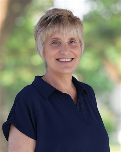 A smiling older woman with short, light blonde hair stands outdoors in front of a softly blurred green background. She is wearing a dark navy blouse with a collar and exudes a friendly, approachable demeanor.